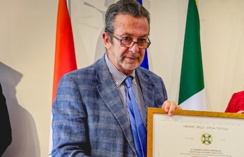 Hombre con barba y gafas, traje a cuadros azul y corbata azul, sostiene un certificado enmarcado de la Orden de la Estrella de Italia. Banderas detrás
