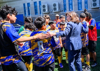 Mujer en traje azul interactúa con jóvenes futbolistas en uniformes azules y amarillos sobre césped verde, con un edificio y unidades de aire acondicionado de fondo