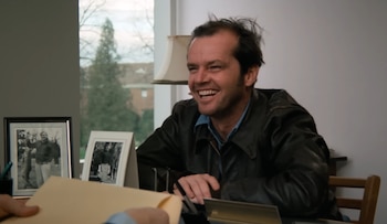 Jack Nicholson con chaqueta de cuero sentado frente a un escritorio con marcos de fotos, una lámpara y un sobre de papel