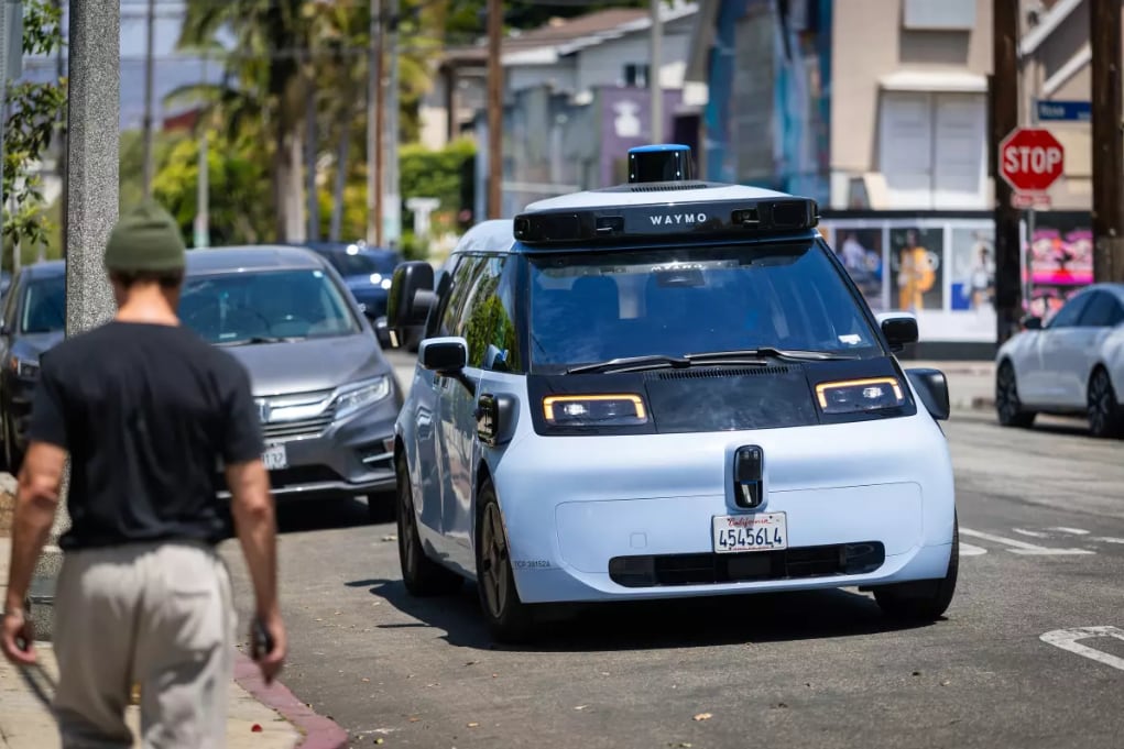 Un vehículo autónomo Waymo, de tipo subcompacto, transita por una calle urbana mientras un peatón camina en primer plano, evidenciando su integración en el paisaje diario de la ciudad. (Allen J. Schaben/Los Angeles Times)