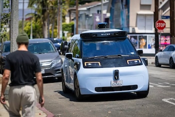 Un coche Waymo autónomo azul claro en una calle urbana. Un peatón camina de espaldas en primer plano. Otros coches y un letrero de 'Stop' son visibles al fondo