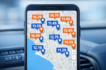 Un smartphone en primer plano muestra una pantalla con un mapa de ciudad. Varios marcadores de precios azules y naranjas con números destacan en el mapa.
