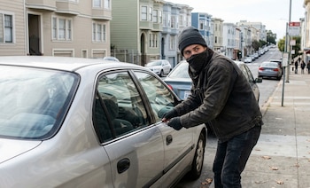 Un hombre encapuchado y con el rostro cubierto intenta abrir la puerta del conductor de un coche plateado estacionado en una calle con casas antiguas