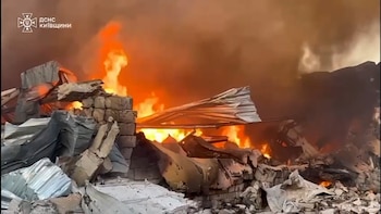 Vista de un edificio en ruinas con grandes llamas naranjas y humo denso elevándose, mostrando escombros y metal retorcido en primer plano