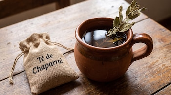Taza de barro con té y hojas secas, bolsa de tela de saco con inscripción "Té de Chaparral", y mesa de madera con vetas visibles.
