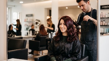 Una mujer de cabello castaño rojizo sonríe mientras un hombre utiliza un secador y un cepillo redondo en un salón de belleza.