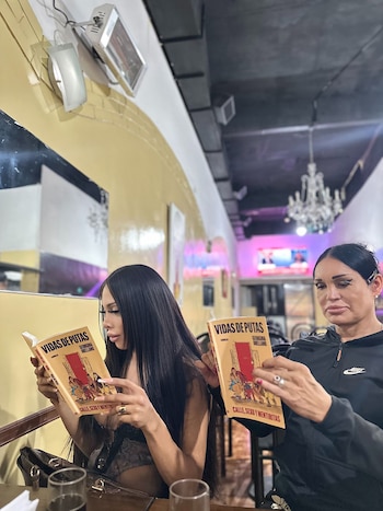 Dos personas sentadas en una mesa de un café. Ambas sostienen y leen un libro de tapa color crema con el título 'Vidas de Putas'. Hay vasos en la mesa