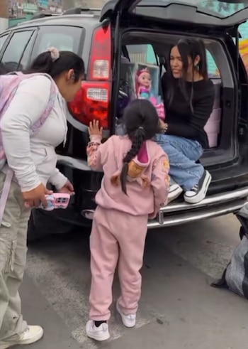 Una mujer de pie a la izquierda, una niña de espaldas con un traje deportivo rosa, y una mujer sentada en el maletero abierto de un automóvil oscuro con una muñeca