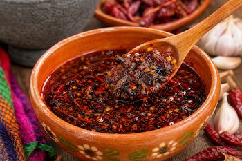 Cuenco de barro con salsa de aceite y semillas, una cuchara de madera, chiles secos rojos, cabezas de ajo y un mortero de piedra.