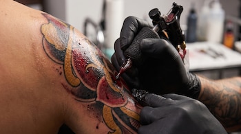 Primer plano de mano enguantada de negro sujetando máquina de tatuar que aplica diseño de tinta negra, roja y amarilla en piel con gotas de tinta y brillo superficial.