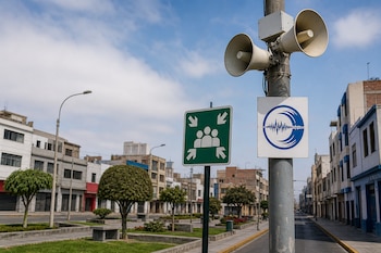 Un poste con dos altavoces blancos, una señal verde de punto de reunión y un cartel con ondas sísmicas junto a una calle con edificios.