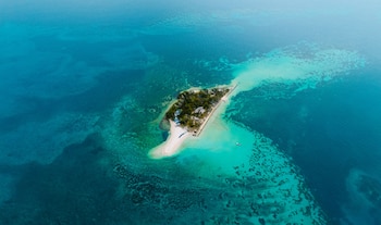 Fotografía aérea de un islote con vegetación, franja de arena blanca y formaciones de arrecife visibles en aguas turquesas y azules