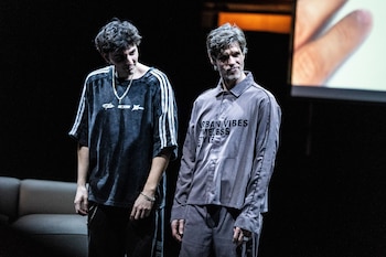 Dos actores en un escenario oscuro. Uno viste camiseta oscura con tiras blancas, el otro camisa gris con texto. Al fondo, una mano proyectada