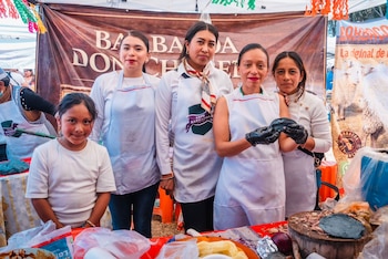 Cinco mujeres, incluyendo una niña, en delantales blancos posan detrás de un puesto de comida en un Marketplace, con masa y platillos sobre la mesa