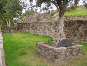 Un paisaje exterior con un área de césped, muros de piedra, un árbol de tronco grueso y un artefacto de piedra oscuro con grabados semicirculares en un recinto de piedra