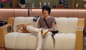 Tommy Lee, el baterista de Mötley Crüe, sentado en un sofá blanco con un perro pequeño y una almohada de gato, con un micrófono delante