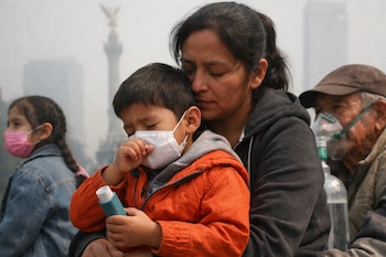 Un niño con mascarilla blanca tose y sostiene un inhalador mientras es abrazado por una mujer; al fondo, un hombre mayor usa una mascarilla de oxígeno.