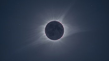La Luna cubre el Sol con una corona blanca y prominencias rojizas en sus bordes, rodeados por un cielo azul grisáceo oscuro con algunas estrellas.