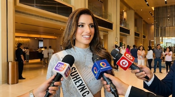 Primer plano de Suheyn Cipriani, de cabello castaño, sonriendo con una banda blanca con 'SUHEYNC' y un vestido plateado, entrevistada por tres micrófonos.