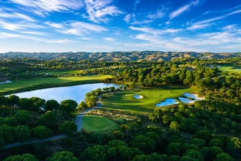 Vista aérea de un campo de golf con césped verde, búnkeres de arena, un lago azul, árboles y colinas al fondo bajo un cielo azul
