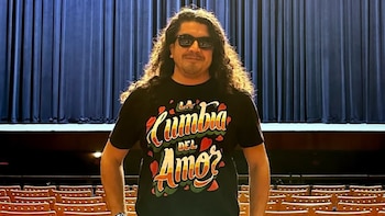 Hombre con cabello rizado y gafas de sol, con camiseta "La Cumbia del Amor", de pie frente a telón azul y sillas de madera