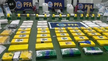 Una mesa de color verde oscuro exhibe numerosos paquetes de droga, principalmente envolturas amarillas con tréboles y blancas, junto a equipamiento de laboratorio. En el fondo se aprecian letreros con el logo de la PFA