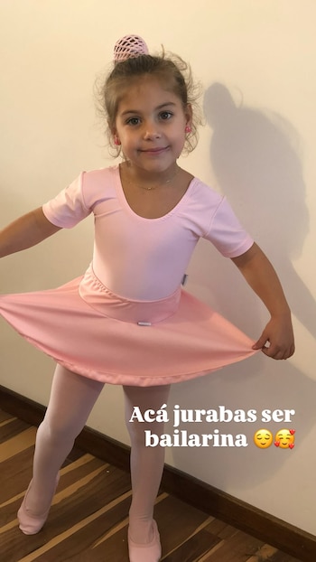 Niña de cabello castaño en moño con red rosa, maillot, falda, medias y zapatillas de ballet rosas, aretes rosas y collar dorado, delante de una pared clara