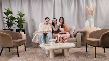 Tres personas sentadas en un sofá beige con cojines, una mesa de centro blanca, dos sillones de terciopelo, dos plantas, un jarrón y cortinas grises
