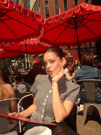 Guille Giles viste una prenda gris y sostiene una carta sentada a la mesa de un restaurante exterior con sombrillas rojas