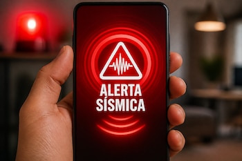 Mano que sostiene un teléfono celular con la frase ALERTA SÍSMICA en pantalla y un icono triangular, con una luz roja al fondo.