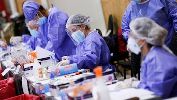 Trabajadores de la salud con batas moradas, mascarillas, protectores faciales y gorros, en un entorno de laboratorio con suministros médicos y botellas