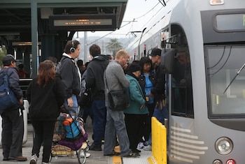 Vista frontal y lateral de un tren gris en una estación con varias personas en el andén, algunas cerca de la puerta del tren y otras esperando