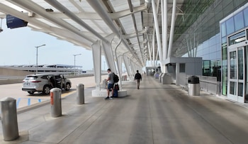 Vista exterior de la entrada de un aeropuerto moderno con una marquesina blanca, una camioneta plateada y personas con equipaje. Se ve un edificio con fachada de cristal