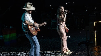 Cantante con traje brillante y micrófono junto a guitarrista con sombrero y pantalones brillantes en el escenario. Confeti, bandera arcoíris y pantalla iluminada visibles