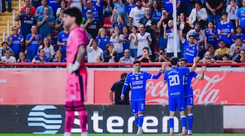 Tres futbolistas vestidos de azul celebran en la cancha mientras la afición aplaude y un portero vestido de rosa figura desenfocado.