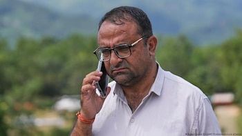 Un hombre con gafas y camisa de manga corta sostiene un teléfono móvil junto a su oído sobre un fondo verde