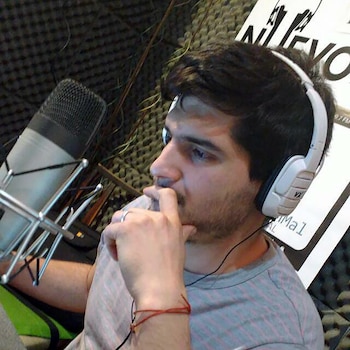 Alejandro Pueblas, un hombre joven de barba, usa auriculares blancos y se sienta frente a un micrófono en un estudio de radio con paneles acústicos