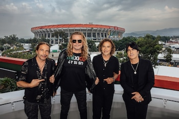 Cuatro hombres de pie en un balcón, uno con camiseta negra que dice "MÉXICO ¿Y SI SÍ?", frente a un estadio, árboles y montañas lejanas