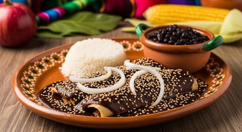 Fotografía de un platón de cerámica con enchiladas de mole oscuro, cubiertas con ajonjolí y aros de cebolla. Acompaña arroz blanco y frijoles negros en cazuela.