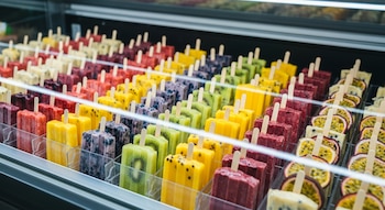 Vista de un congelador de supermercado lleno de filas de paletas heladas artesanales de distintos colores y sabores frutales, incluyendo kiwi y maracuyá.