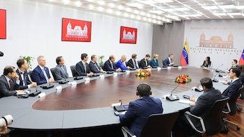 Delegados en trajes sentados en una mesa ovalada de conferencias con micrófonos. La bandera de Venezuela y logos del Palacio de Miraflores se ven en la pared