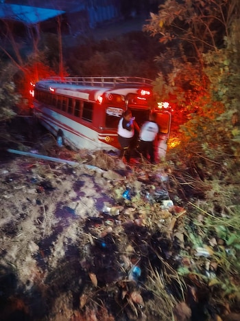 Autobús rojo y blanco accidentado fuera de la carretera en una zona boscosa. Dos personas con uniformes de emergencia están cerca del vehículo