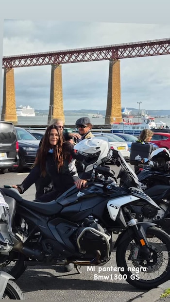 Carolina Baldini posa junto a una motocicleta negra en un estacionamiento, con personas y un puente metálico rojo al fondo