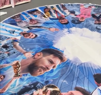 Una falda de tela circular extendida en el piso con retratos de Lionel Messi, en una habitación con percheros de ropa y un sofá