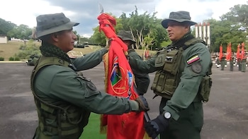 Dos soldados con uniforme verde, sombreros y chalecos tácticos sostienen un estandarte rojo en un patio al aire libre