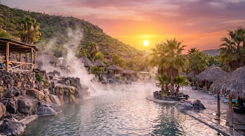 Aguas termales con vapor, palmeras, chozas de paja, rocas y montañas. Sol amarillo en cielo anaranjado de atardecer.