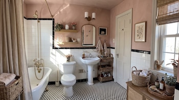 Un baño con bañera, inodoro, lavabo de pedestal, espejo, estantes de madera, cestas de mimbre, paredes rosas y suelo de mosaico blanco y negro.
