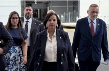 Una mujer con traje azul oscuro y camisa blanca en el centro, un hombre con traje azul oscuro y corbata roja a la derecha, y una mujer con gafas, camiseta azul y falda estampada a la izquierda. Hay otras personas parcialmente visibles. El fondo es un edificio