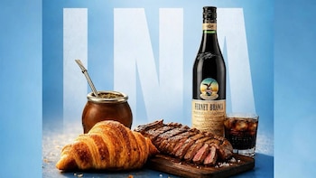 Una botella de Fernet Branca, un mate con bombilla, un croissant, carne asada en tabla y un vaso de Fernet con cola, sobre fondo azul claro