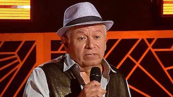 Un hombre mayor, con sombrero fedora a rayas y chaleco oscuro, sostiene un micrófono en un escenario con fondo negro y diseños geométricos naranjas